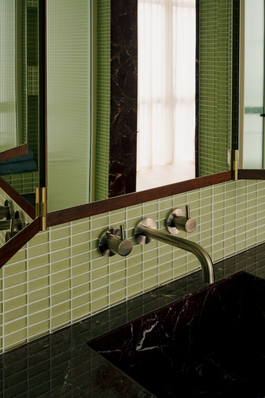 Wall-mounted taps in brushed brass complement the gridded mosaic tiles, with the burgundy marble providing a warm contrast to the cool green tones of the private spaces