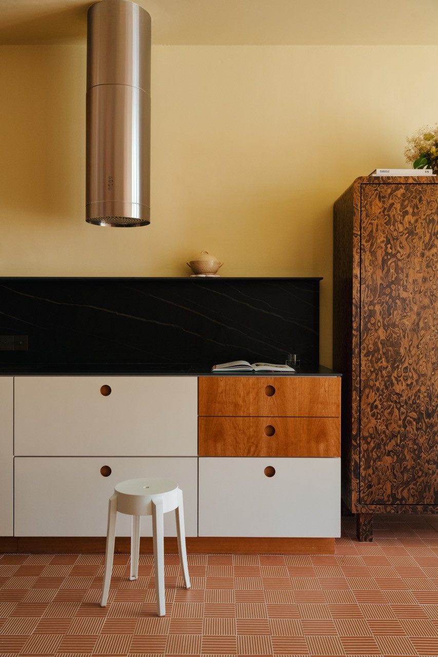 The California burl veneer cabinet stands prominently beside the kitchen, its rich wood grain contrasting with the clean lines of the white and cherry wood kitchen cabinetry and dark sintered stone surfaces