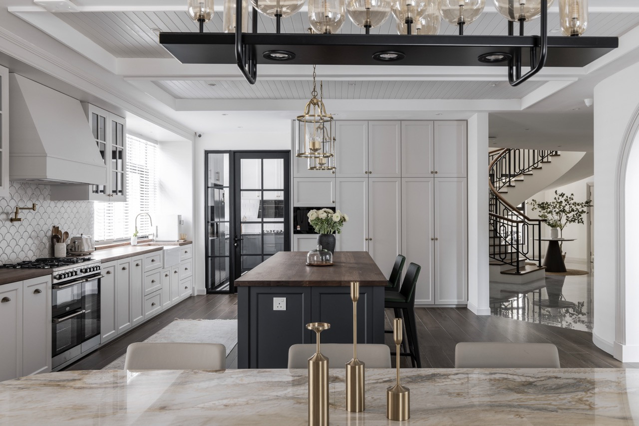 The kitchen showcases painted beam ceilings that reference farmhouse traditions, while Shaker-style cabinetry in soft grey pairs with a dark island base