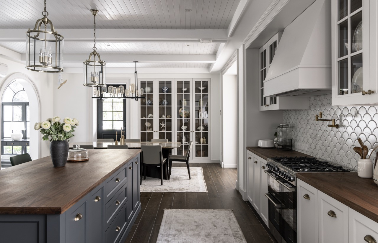 The kitchen showcases painted beam ceilings that reference farmhouse traditions, while Shaker-style cabinetry in soft grey pairs with a dark island base