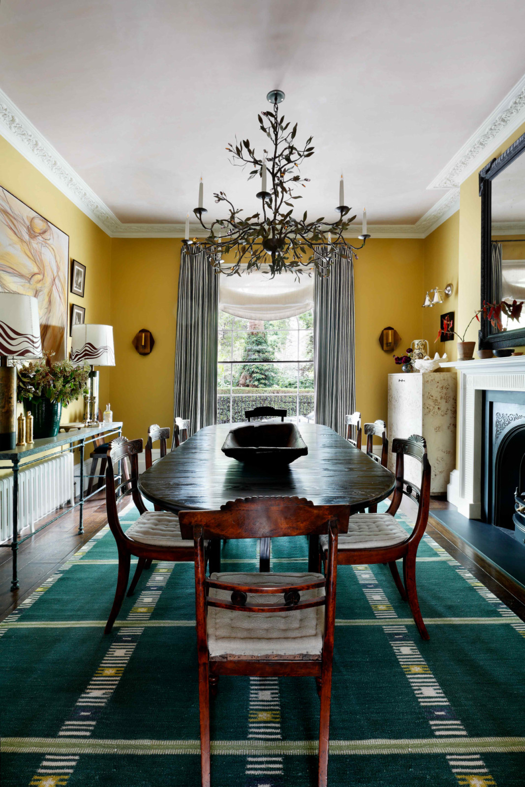 An ornate Cox London chandelier suspends above a worn ebony dining table and antique dining chairs