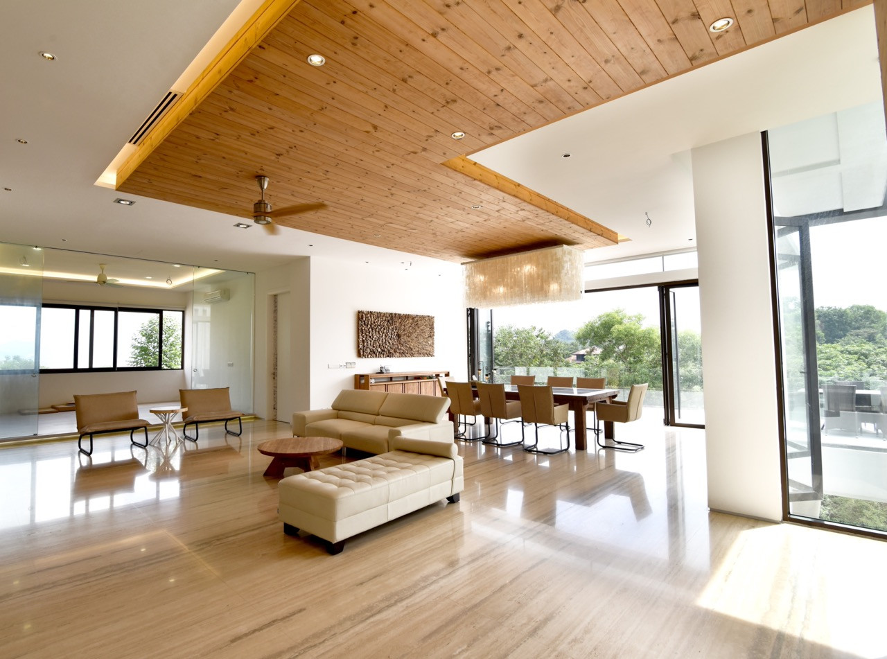 The dining area looks out through floor-to-ceiling windows to the surrounding greenery, with timber ceiling elements providing warmth against white walls