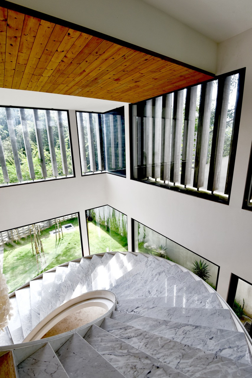 The curved marble staircase winds upward through floor-to-ceiling glazing that offers views down to the internal courtyard and surrounding forest