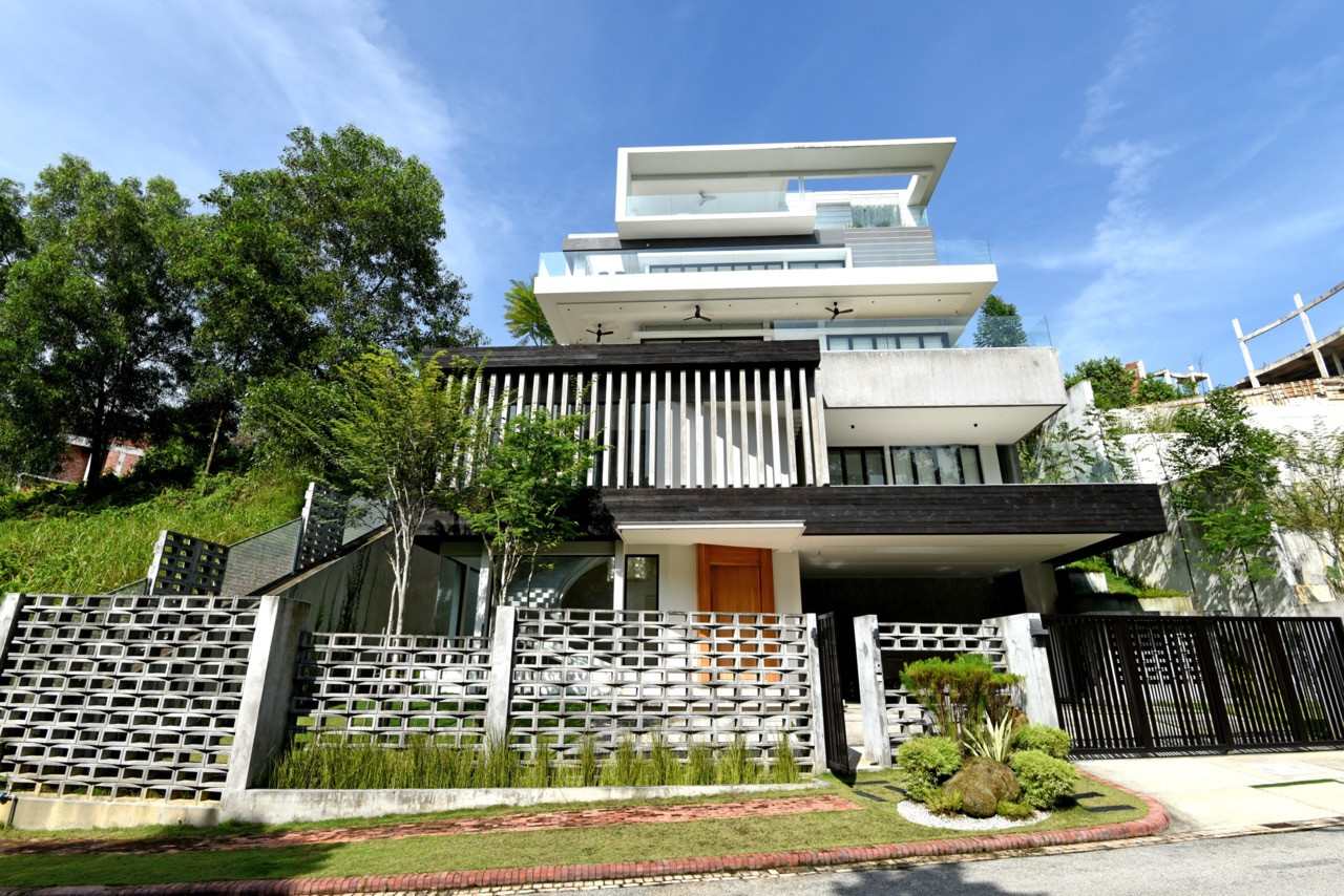 Street view of the hillside home showing the stacked concrete volumes with timber screening elements, perforated boundary walls, and the natural slope rising behind the property