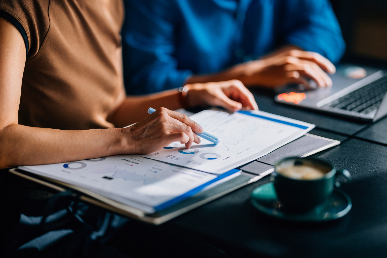 Two colleagues reviewing financial data and charts. The focus is on teamwork and analytical skills in a modern office setting with a laptop and coffee. (ภาพ: FreshSplash via Getty Images)