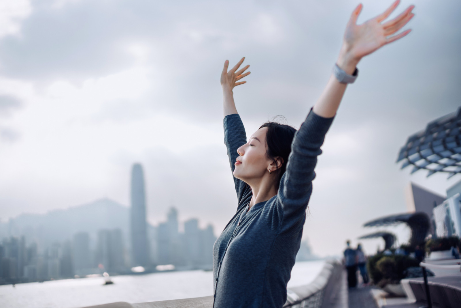 Beautiful young Asian woman with her eyes closed, taking a deep breath and stretching arms against Victoria Harbour and urban city skyline