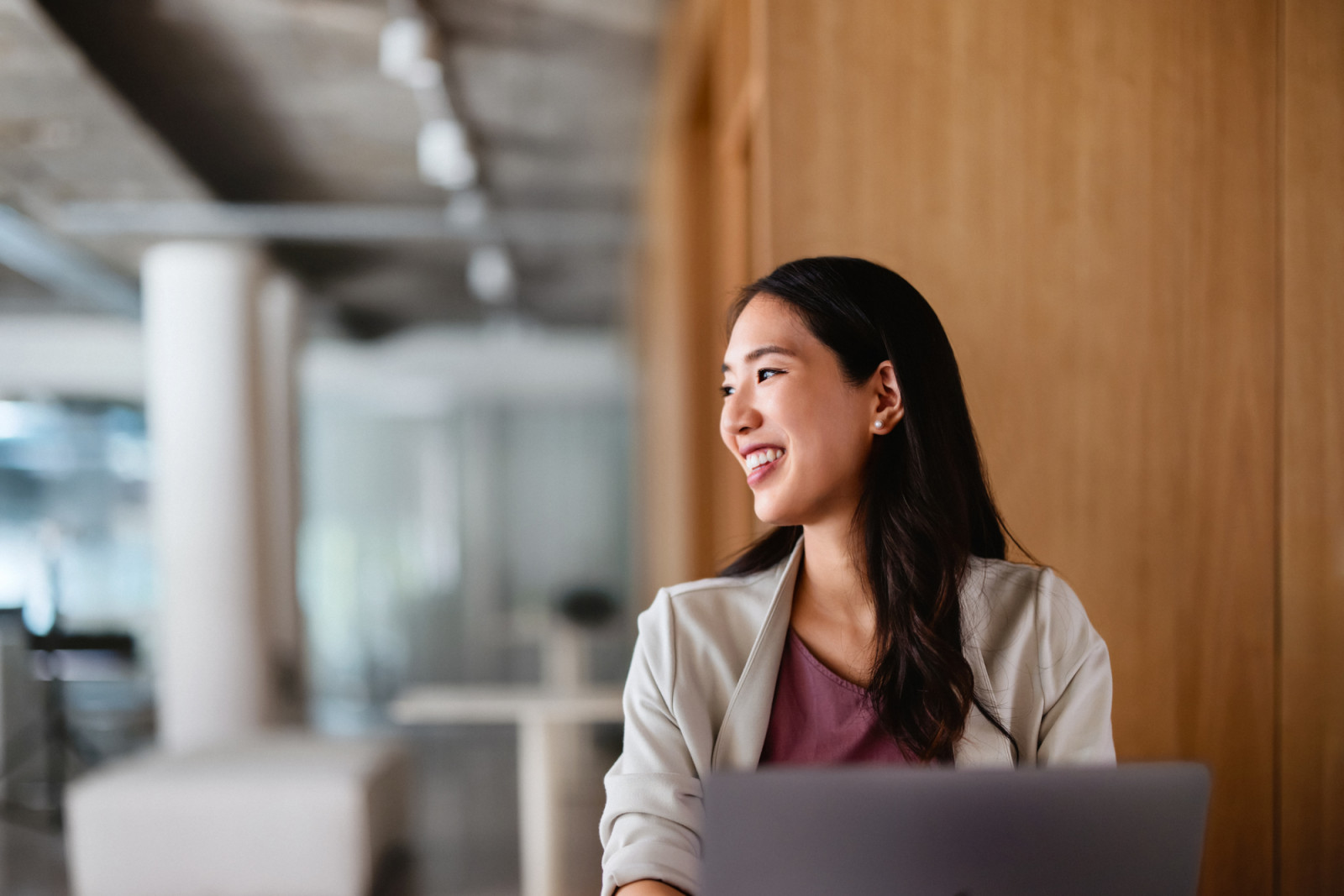 Confident young Asian businesswoman smiling while sitting at a desk in a modern office setting, exuding positivity and success.