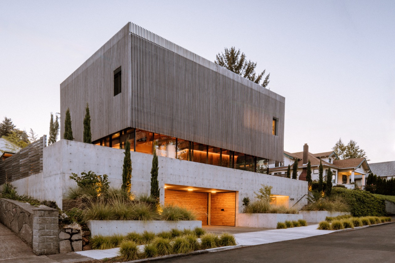 The home sits elevated above street level with concrete retaining walls, native landscaping by Lilyvilla Gardens, and a wood garage door