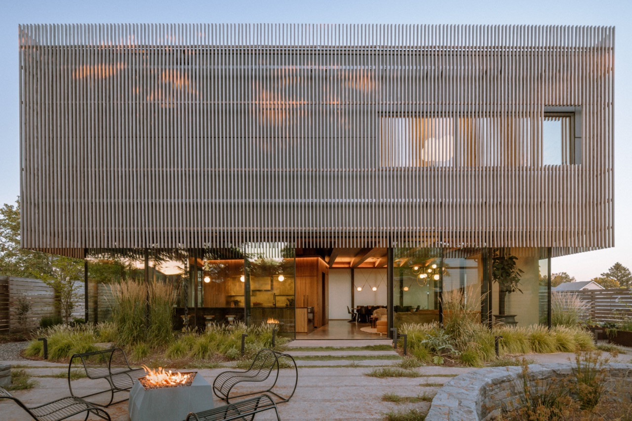 The evening exterior reveals the thermally modified pine cladding that will weather from caramel to silver brown, with the glass-wrapped main level glowing from within
