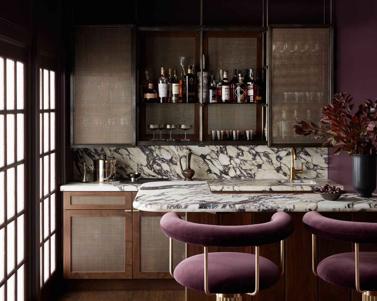 The plum-toned bar, designed in collaboration with architect Neil Young, features laterally moving mesh metal shelves and dramatic marble countertops, complemented by velvet bar stools by Thomas Hayes, which pair well with the Farrow & Ball Brinjal walls.