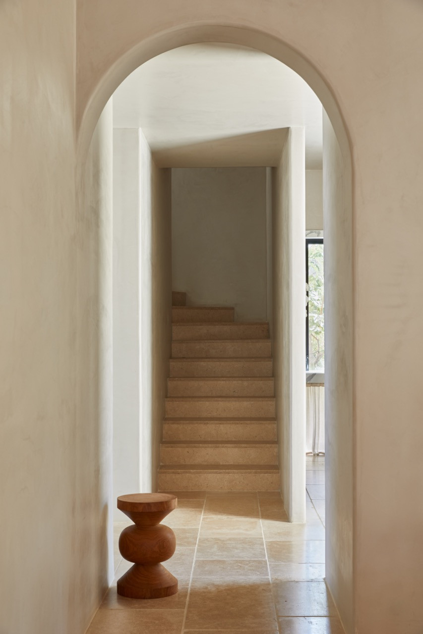 An arched doorway finished in patiti plaster frames a view to wooden stairs, with a sculptural side table in the foreground