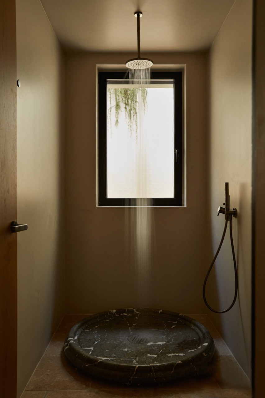 A hand-carved dark marble shower basin sits beneath a rainfall fixture in a minimalist bathroom where the window frames nature as the primary focal point