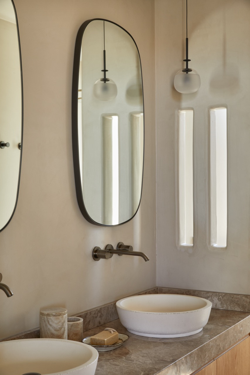 An oval mirror with black metal frame hangs above dual vessel sinks on a marble countertop, flanked by frosted glass pendant lights