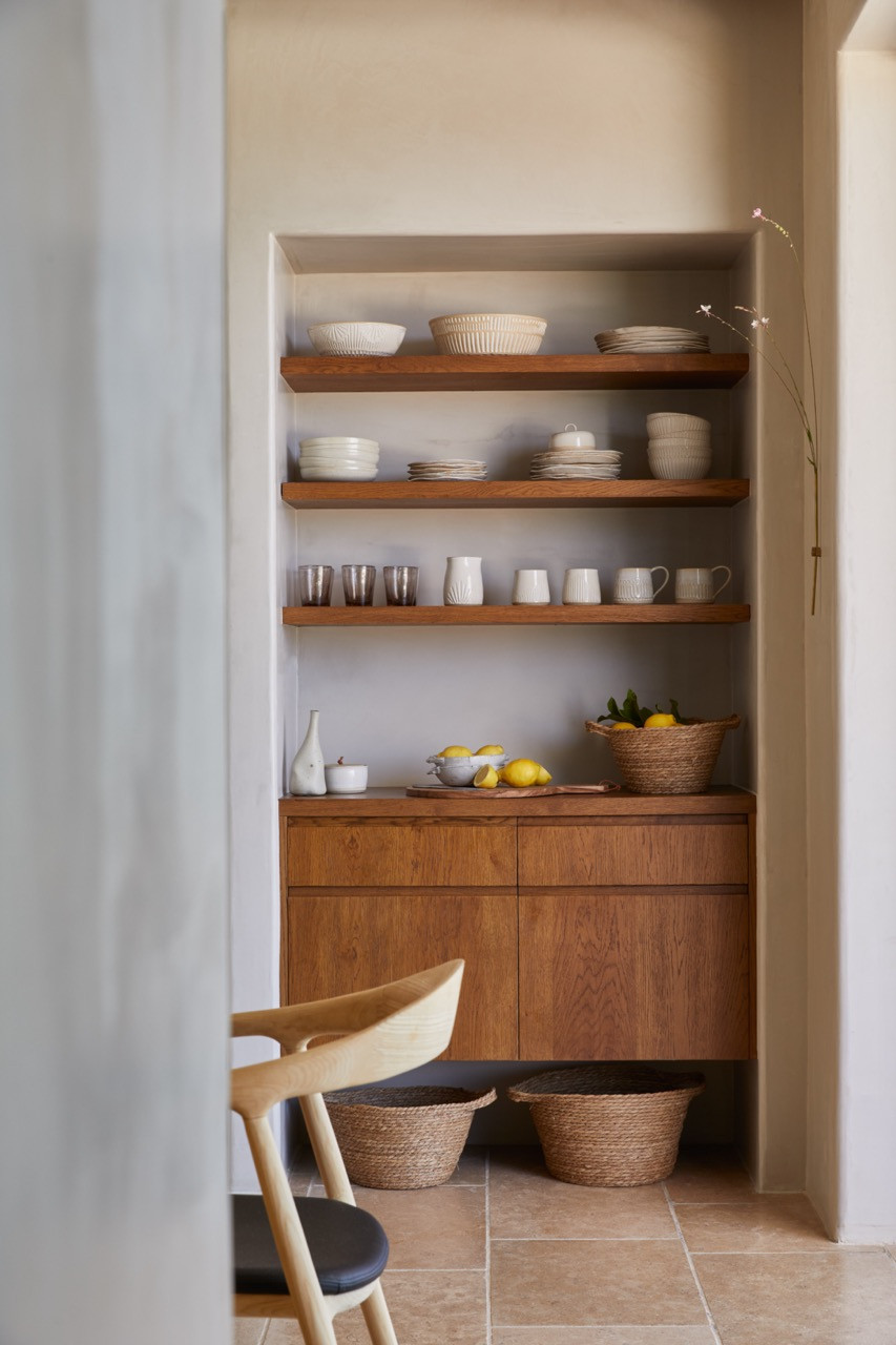 A built-in pantry niche features wooden shelving displaying ceramics and everyday items above wooden cabinetry with woven basket storage