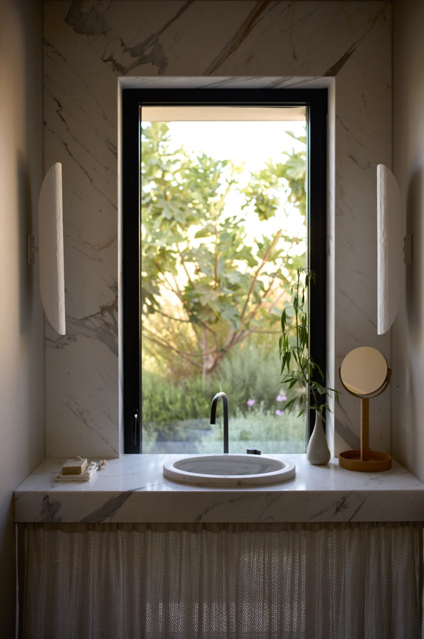 A marble-topped vanity sits before a window framing views of the garden, flanked by vertical sconces