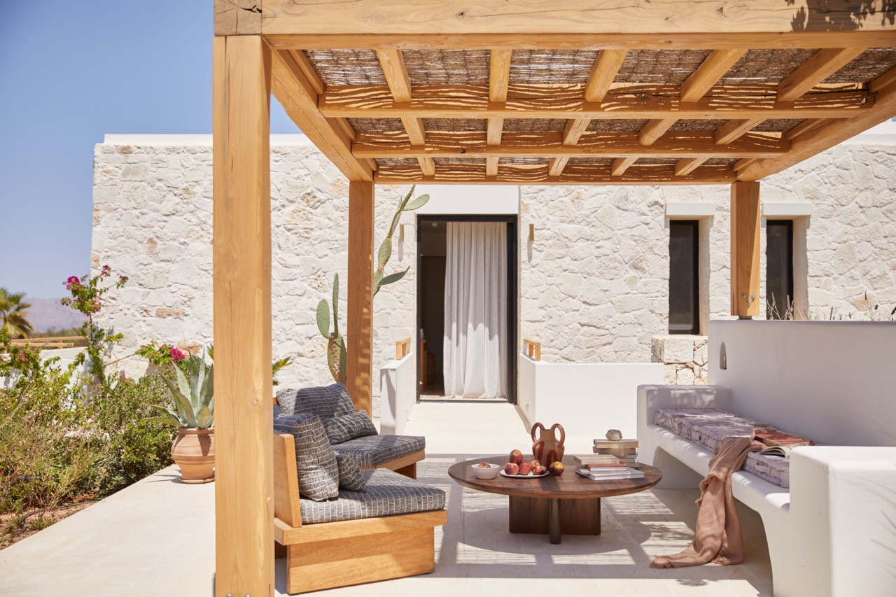 A wooden pergola with natural reed screening provides shade for the outdoor seating area featuring custom furniture against white stone walls