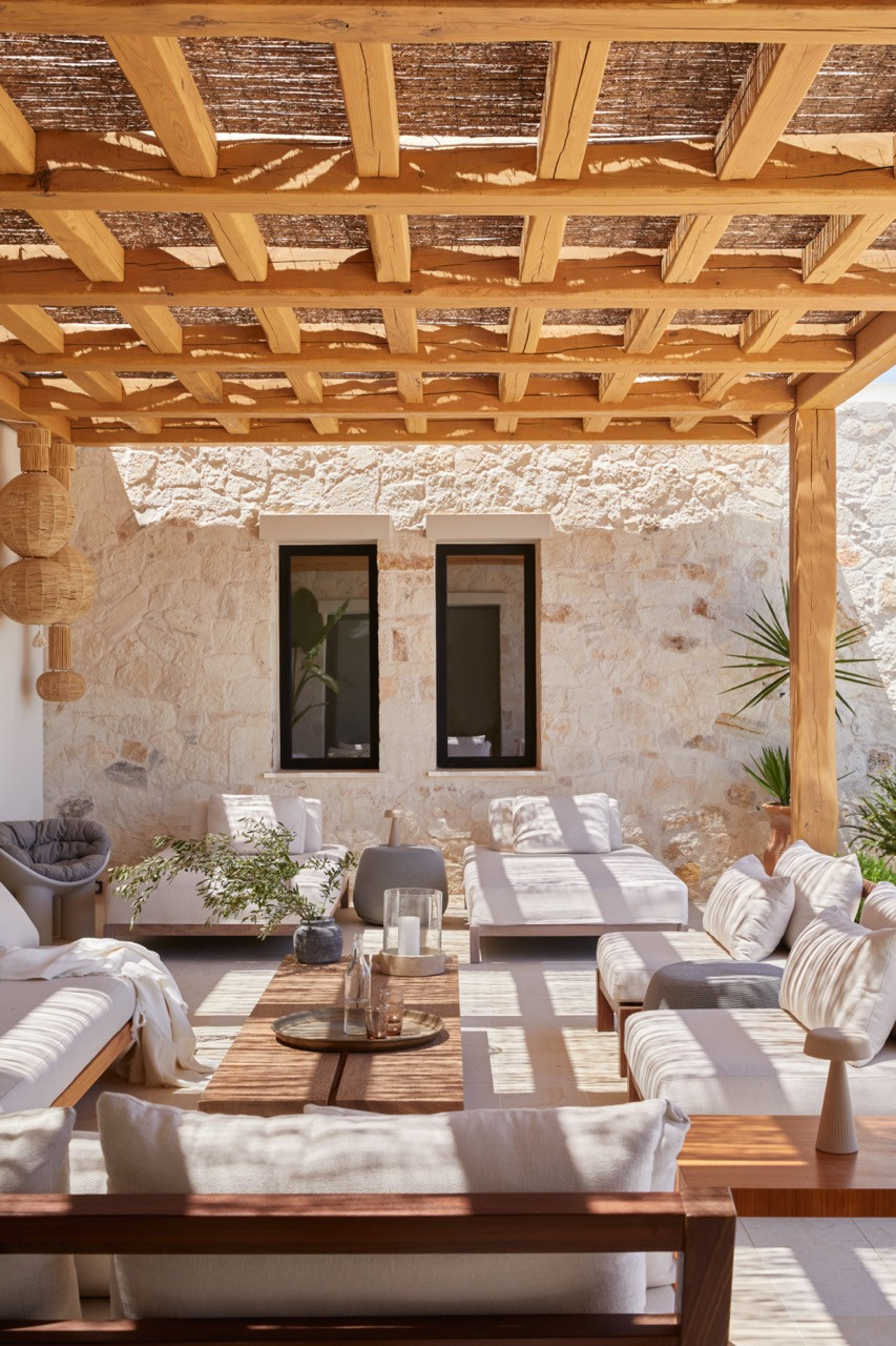 Custom outdoor furniture from Cari Giannoulias Design sits beneath a hand-cut wooden pergola against walls finished in hand-chiseled stone