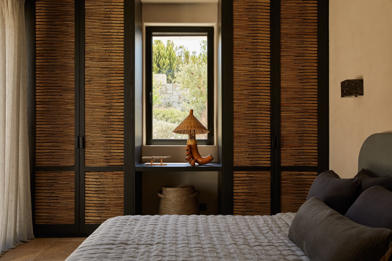 The bedroom features floor-to-ceiling wardrobes with textured rattan panels flanking a window that overlooks the Mediterranean landscape
