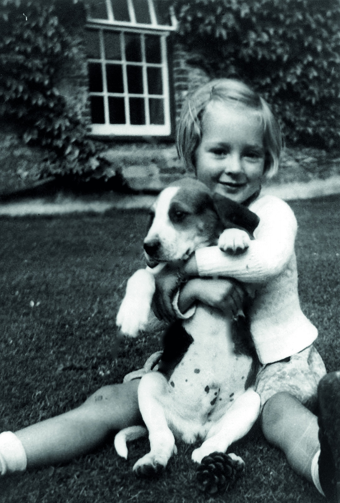 Conservationist Jane Goodall in her childhood days, seated in a garden in England, and she is hugging a puppy. The photo is in black & white
