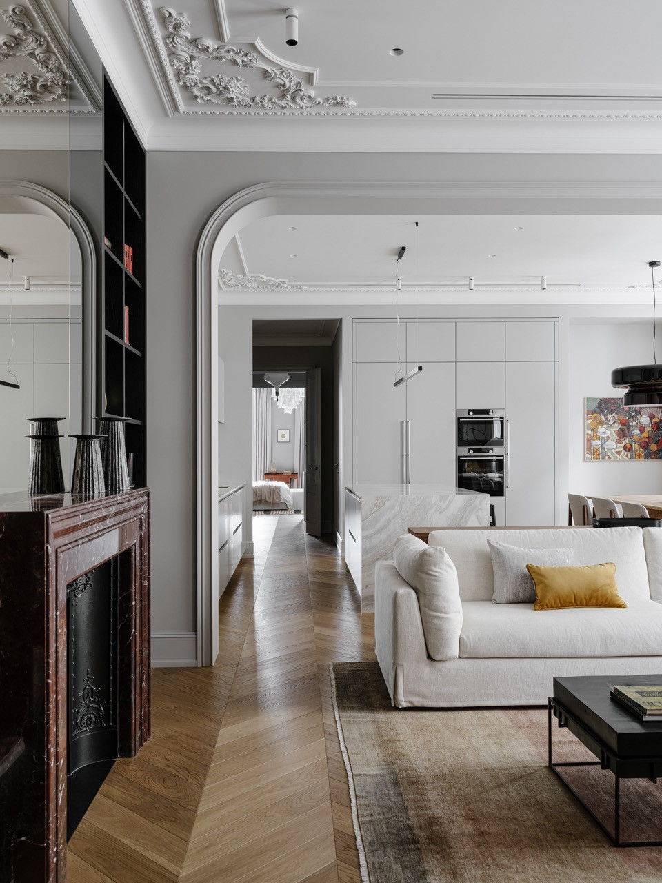 View from library through arched opening to living area with custom dark shelving, marble fireplace and herringbone parquet throughout