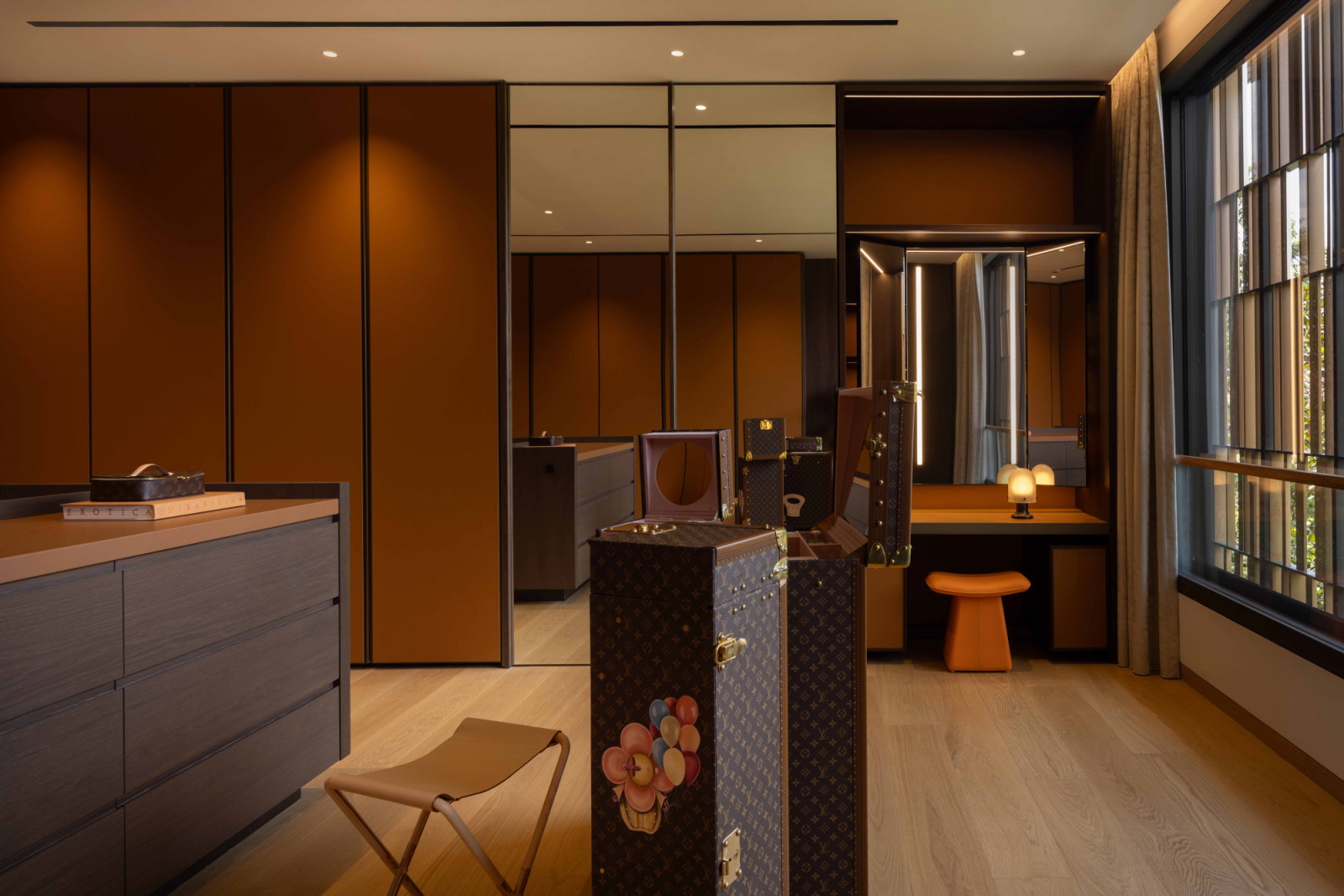 The ultra-stylish walk-in wardrobe features dark wood and leather finishes that evoke a luxury fashion boutique while Louis Vuitton trunks are arranged almost as sculptures