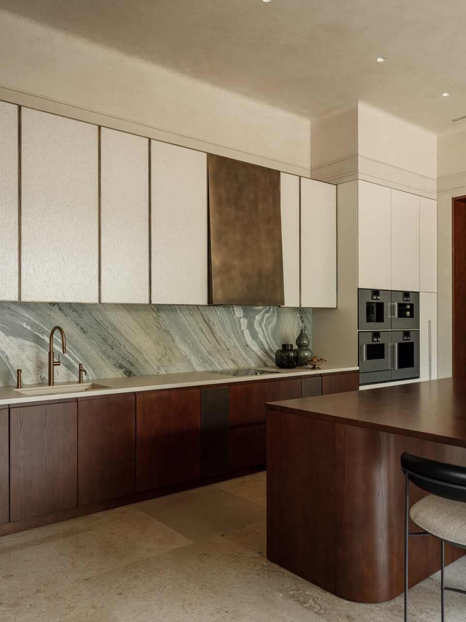Kitchen view showing full run of cabinetry with white uppers and warm wood lowers, book-matched marble backsplash, curved island, and integrated appliances