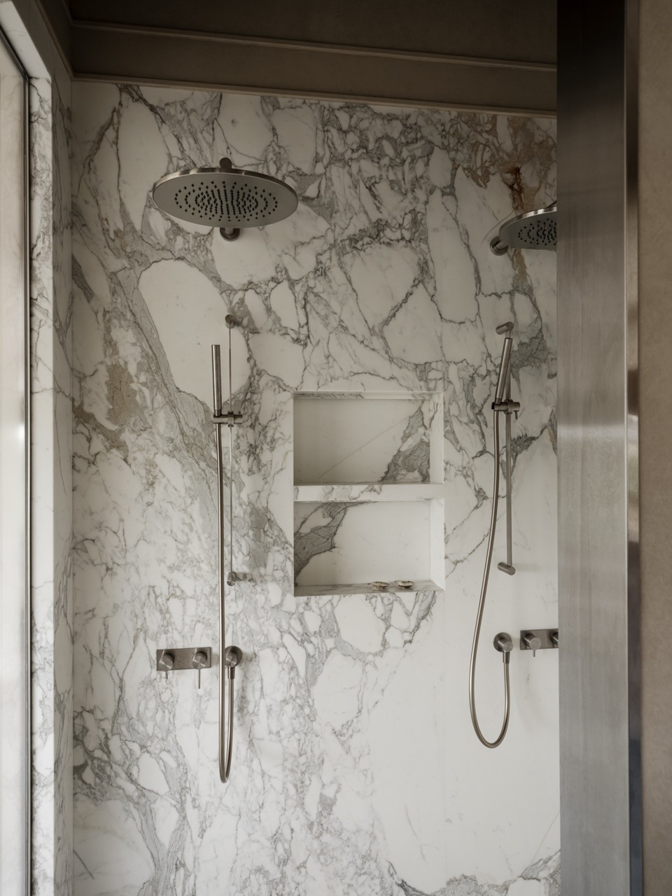 Master bathroom shower with Calacatta Vagli marble walls showing dramatic veining, dual rainfall showerheads, and integrated storage niche