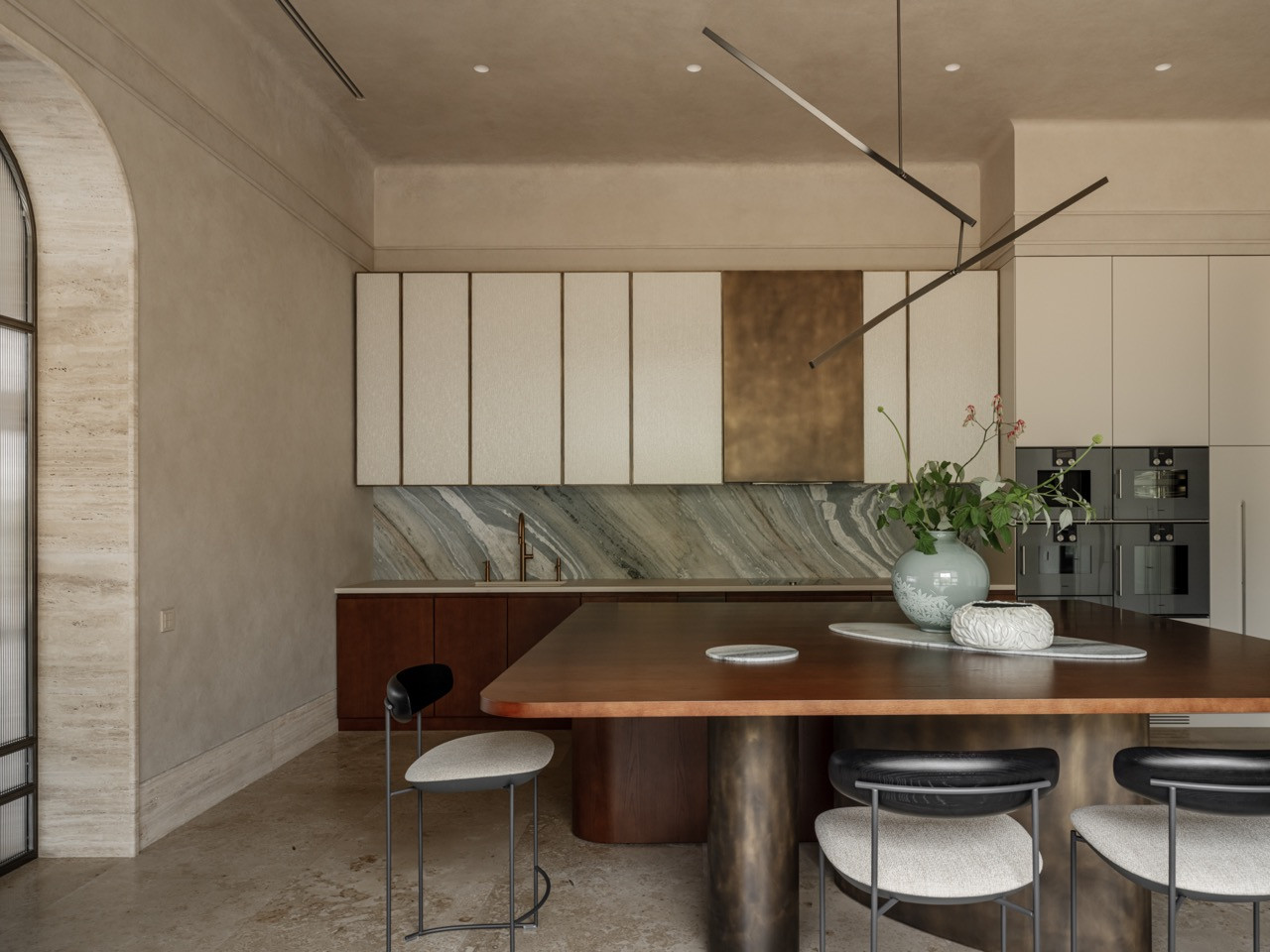 Kitchen featuring white upper cabinets with brass accent, book-matched marble backsplash, curved island with brass cylindrical base, and bar stools by Modenature