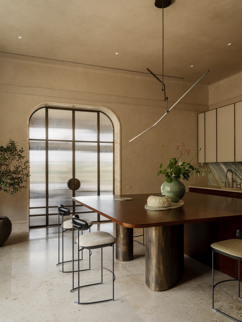 Kitchen-dining area showing arched glass partition, potted olive tree, curved dining table with brass legs, and bar stools by Modenature at the island
