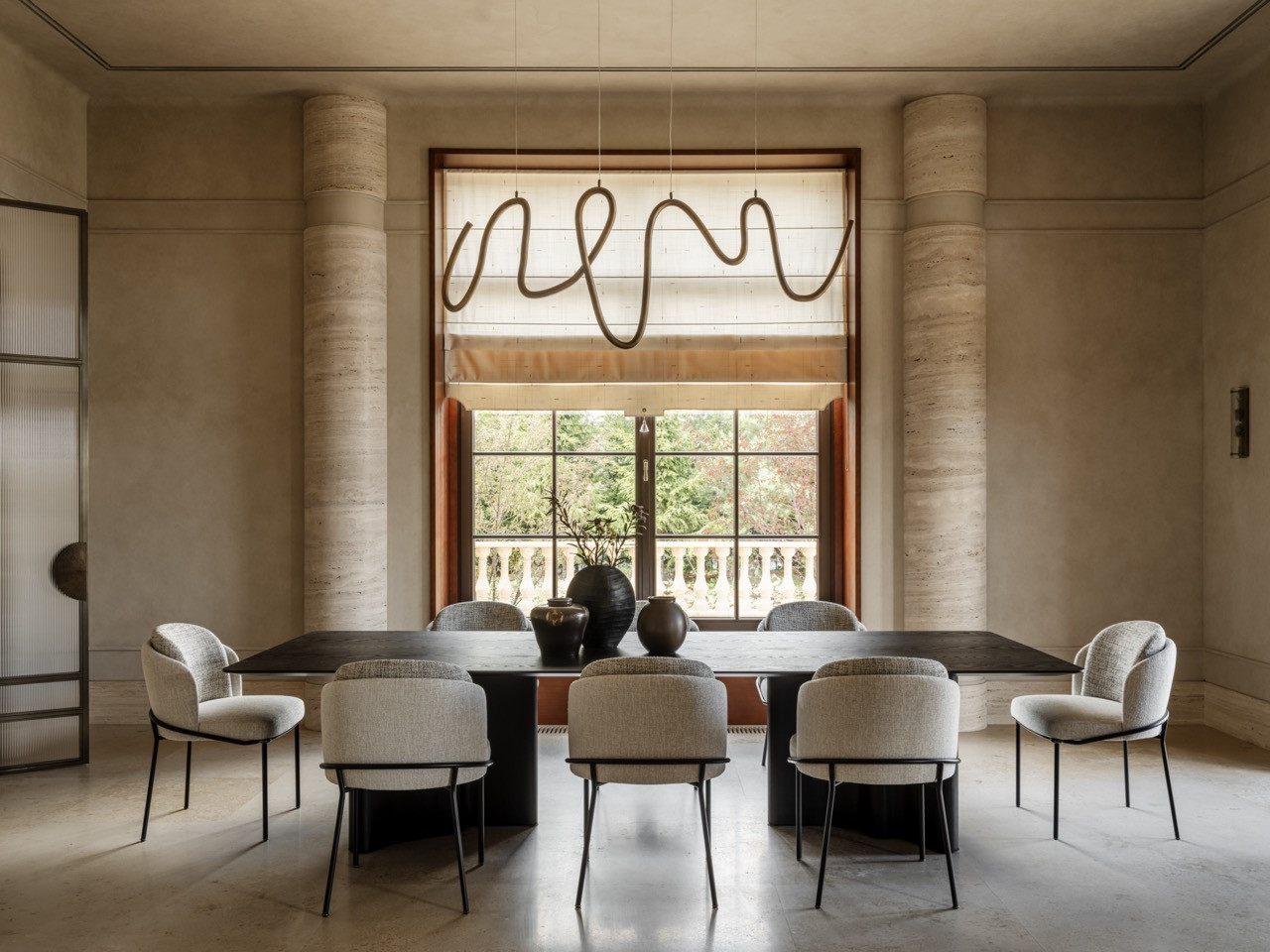 Dining room with travertine columns, oval table by Point Woodcraft in burr oak veneer, Modenature chairs, sculptural brass pendant by Ango, and plaster moulding details