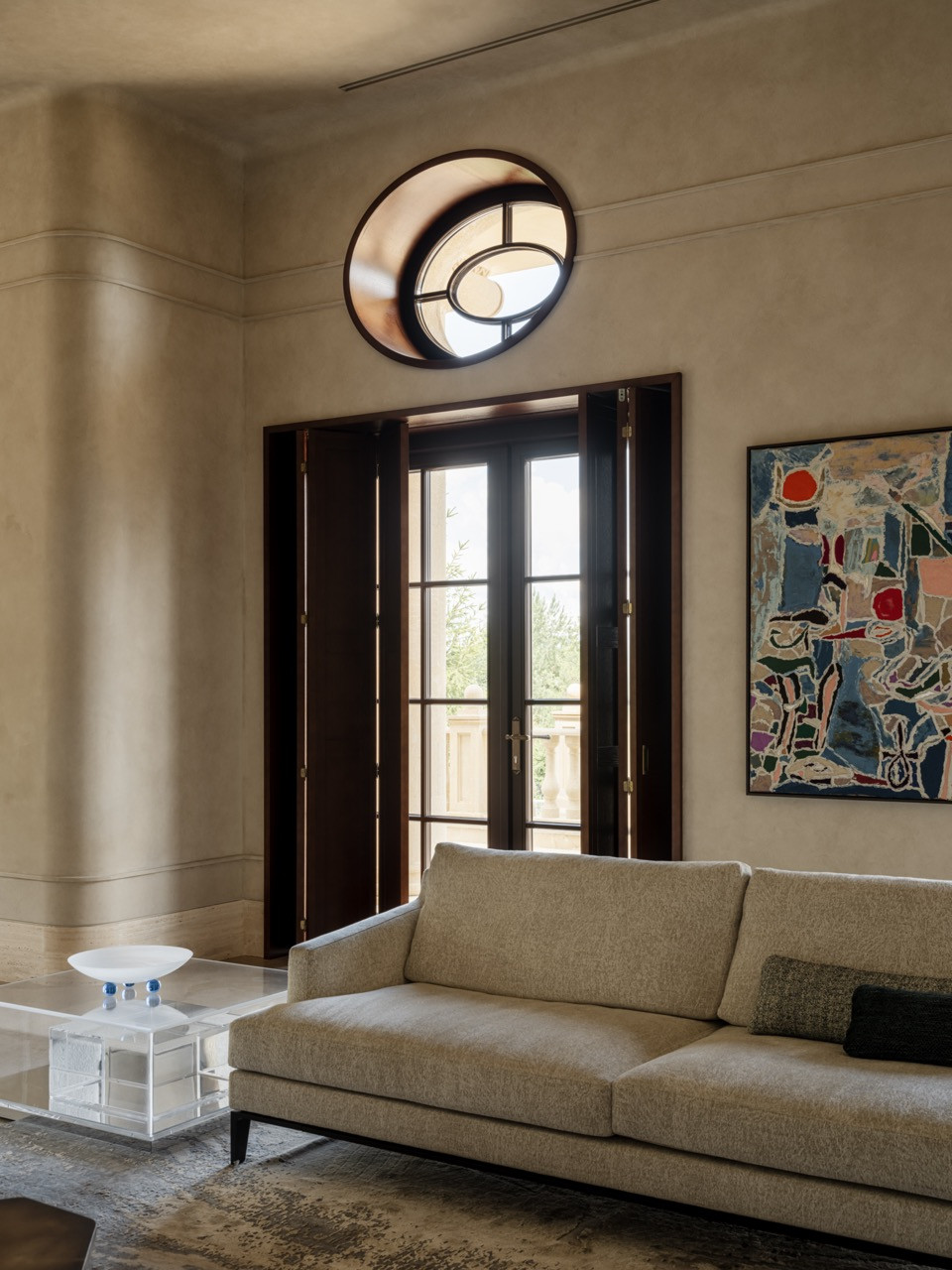 Living room detail showing Modenature sofa, acrylic coffee table with blue glass accents, circular wall mirror, and abstract artwork with French doors to balcony beyond