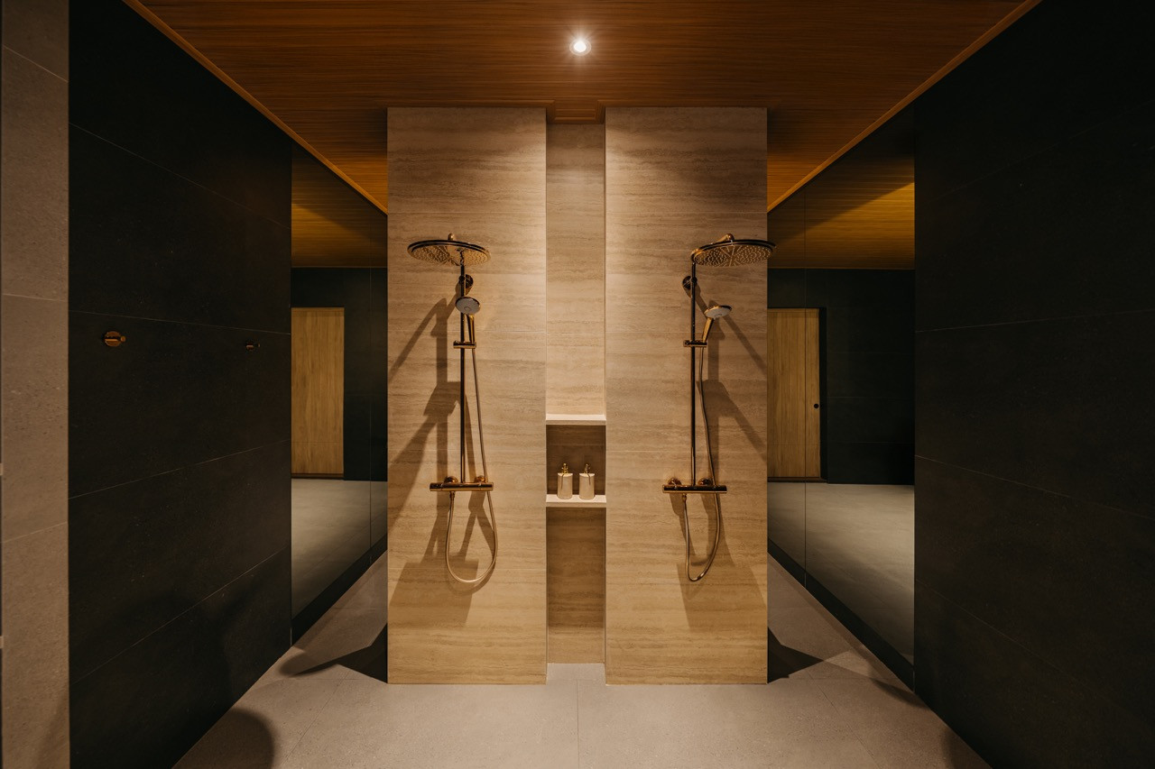Twin shower areas with brass fixtures and timber ceiling panels, the bathroom follows the condominium’s material palette of dark stone and warm timber