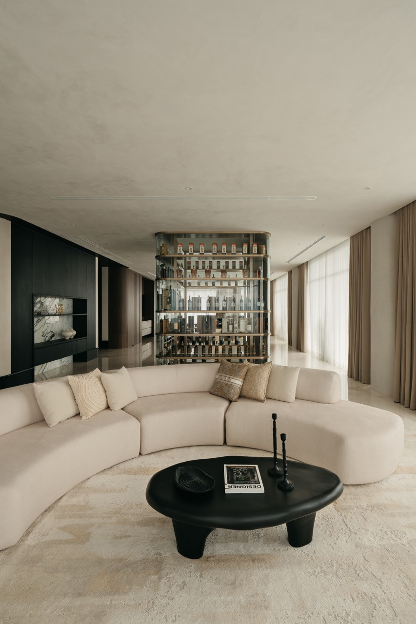 A curved sofa sits in the living area with the backlit spirits cabinet visible in the distance, and floor-to-ceiling sheer curtains line the exterior walls