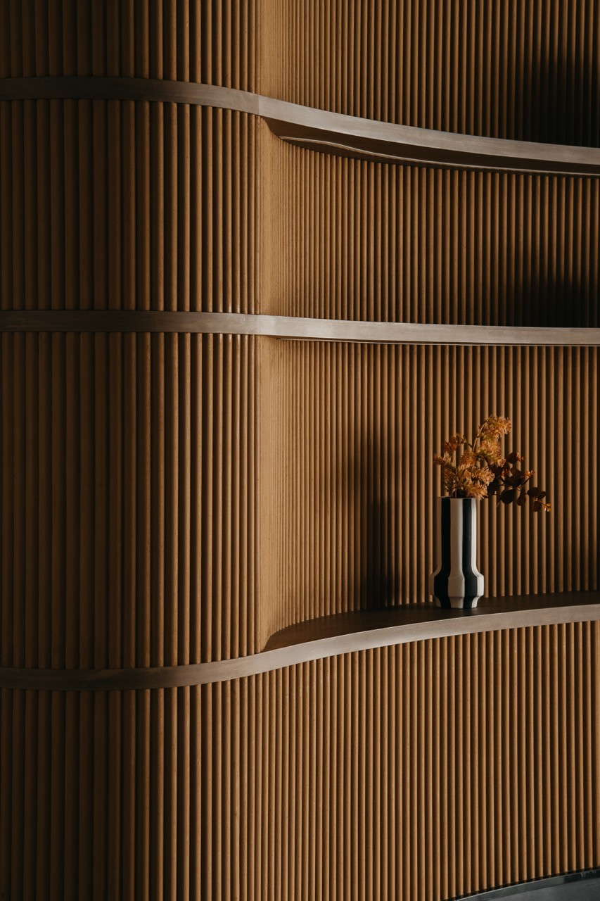 A detail of the curved timber shelving shows the ribbed backing and floating display surfaces with objects on the backlit shelves
