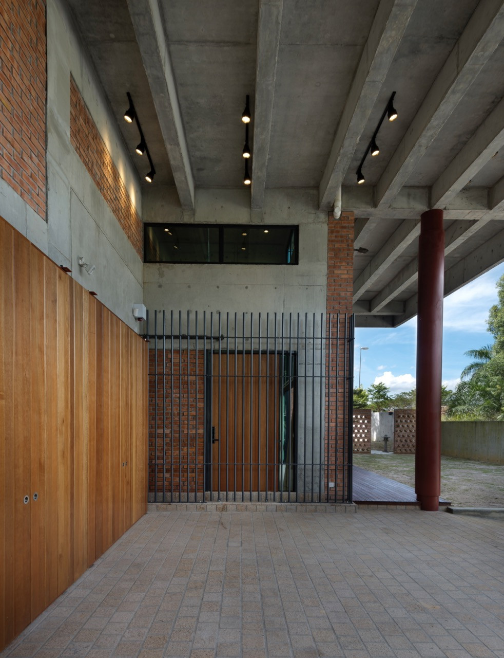 The material palette layers across the covered arrival space: timber garage doors, perforated brick screen walls, raw concrete structure and steel columns
