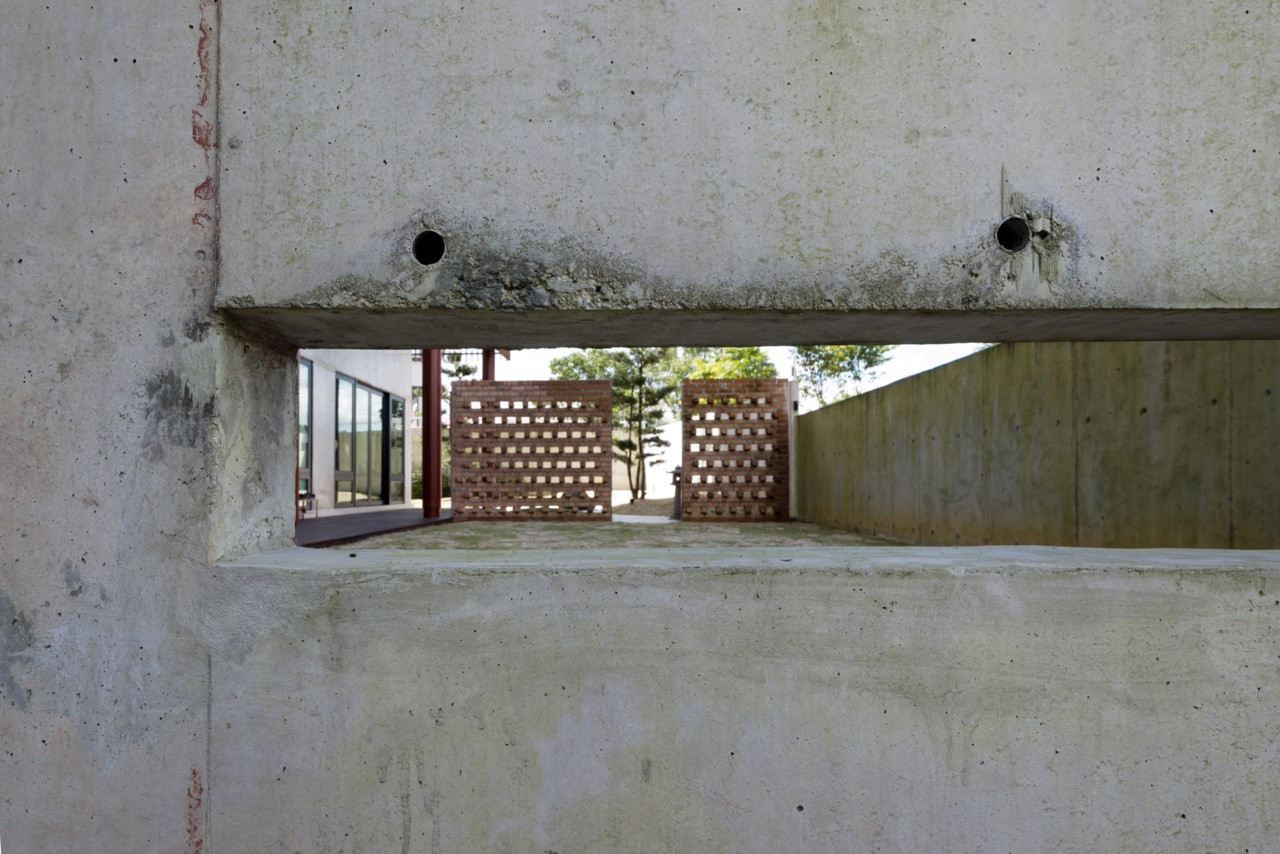A rectangular opening in the raw concrete wall frames views through to the perforated brick screen walls and garden beyond