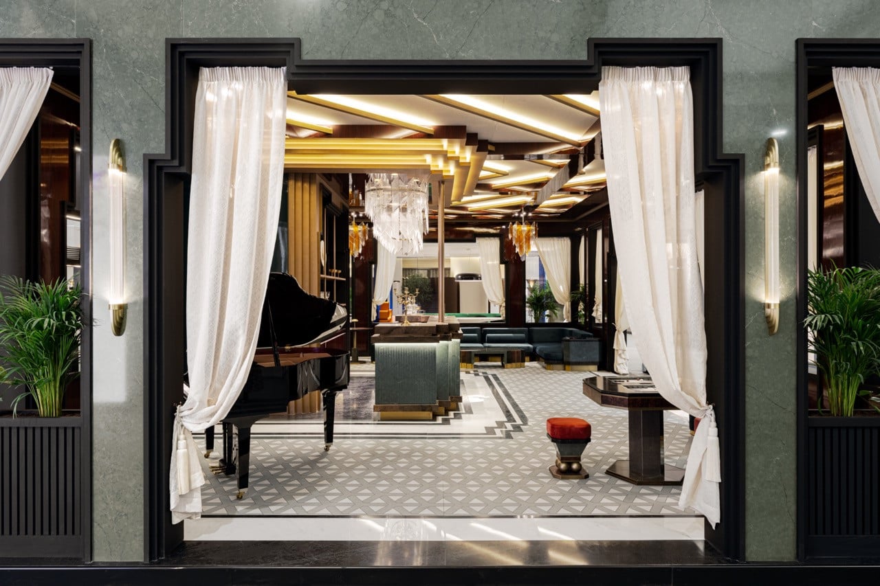 Looking through the entrance portico clad in grey stone-look Cosentino surfaces, the view encompasses a grand piano, game tables, lounge seating and the bar, with the geometric floor pattern in engineered stone creating visual continuity across the interconnected spaces