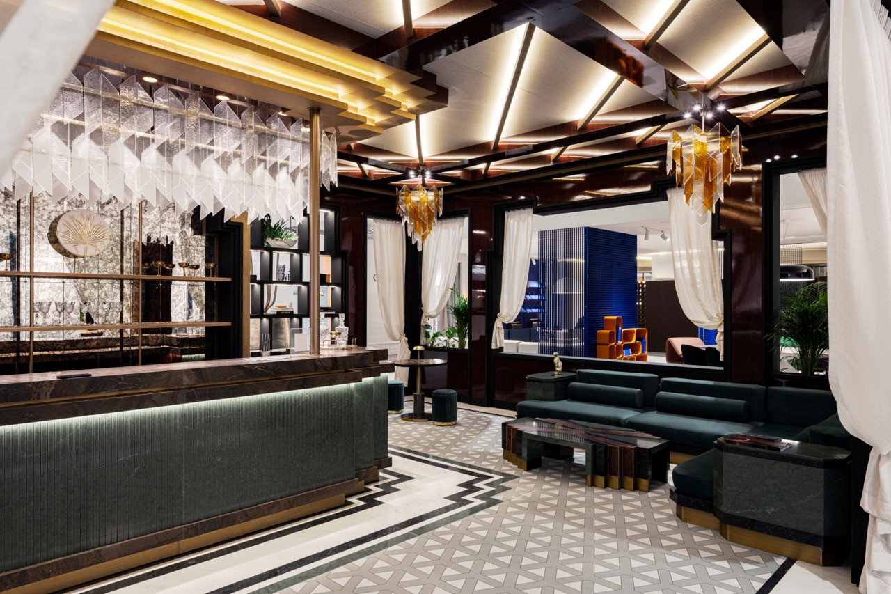 The full lounge interior reveals multiple zones, including the green fluted bar, teal velvet seating, geometric chandeliers, and a stepped ceiling detail, with all horizontal and vertical surfaces fabricated from Cosentino’s Silestone, Dekton and Sensa ranges in jewel tones and dark neutrals