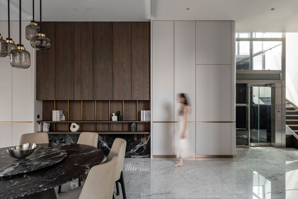 The dining area features clustered pendant lights suspended above a dark marble table, with open shelving displaying vertical brass trims and maintaining sight lines to the living space and upper circulation