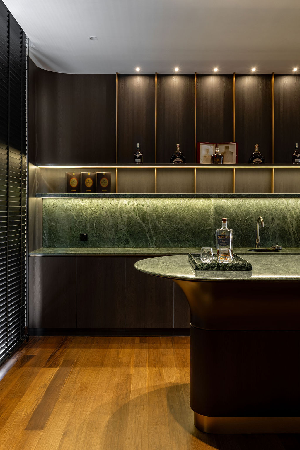 The entertainment room's wet bar integrates green-veined marble as a backsplash with dark wooden joinery featuring bronze trims and open shelving with vertical detailing