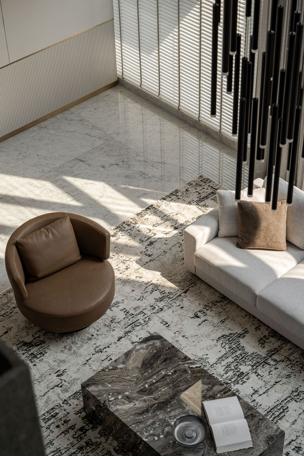 The double-height living space features a flamed-granite-textured wall against a soaring ceiling, with vertical ridges on dark wood accents framing the suspended linear chandelier above a seating arrangement anchored by a green-veined marble coffee table