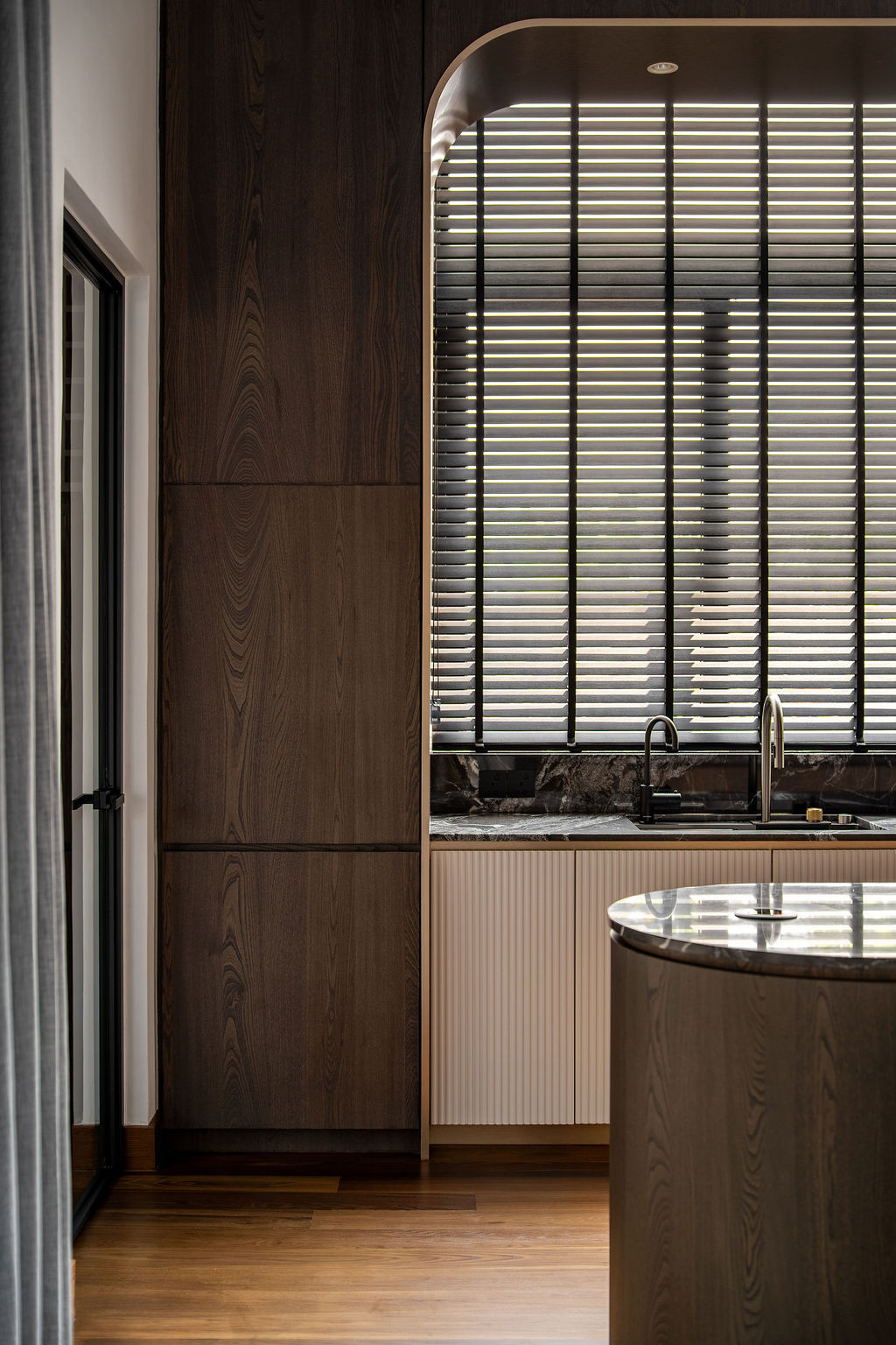 The curved fluted glass arched door frames the entertainment room’s wet bar beyond, with dark wood panelling and vertical detailing reinforcing the linear rhythm established throughout the residence