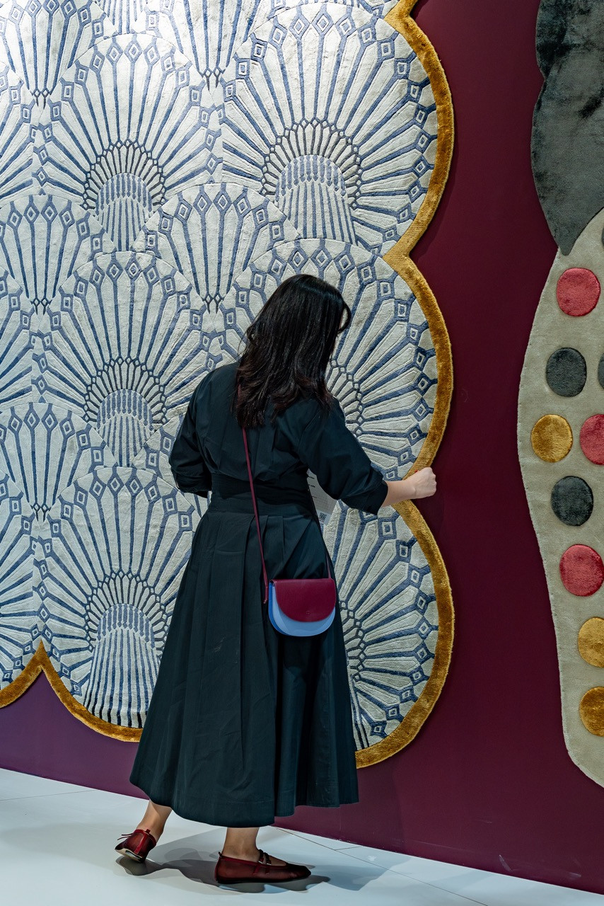 A visitor examines Khazzanz, the rug inspired by Dubai’s Al Khazzan Water Tower, featuring radiating blue and white patterns with gold trim from the Postcards collection by HANDS Carpets and Doodle And The Gang