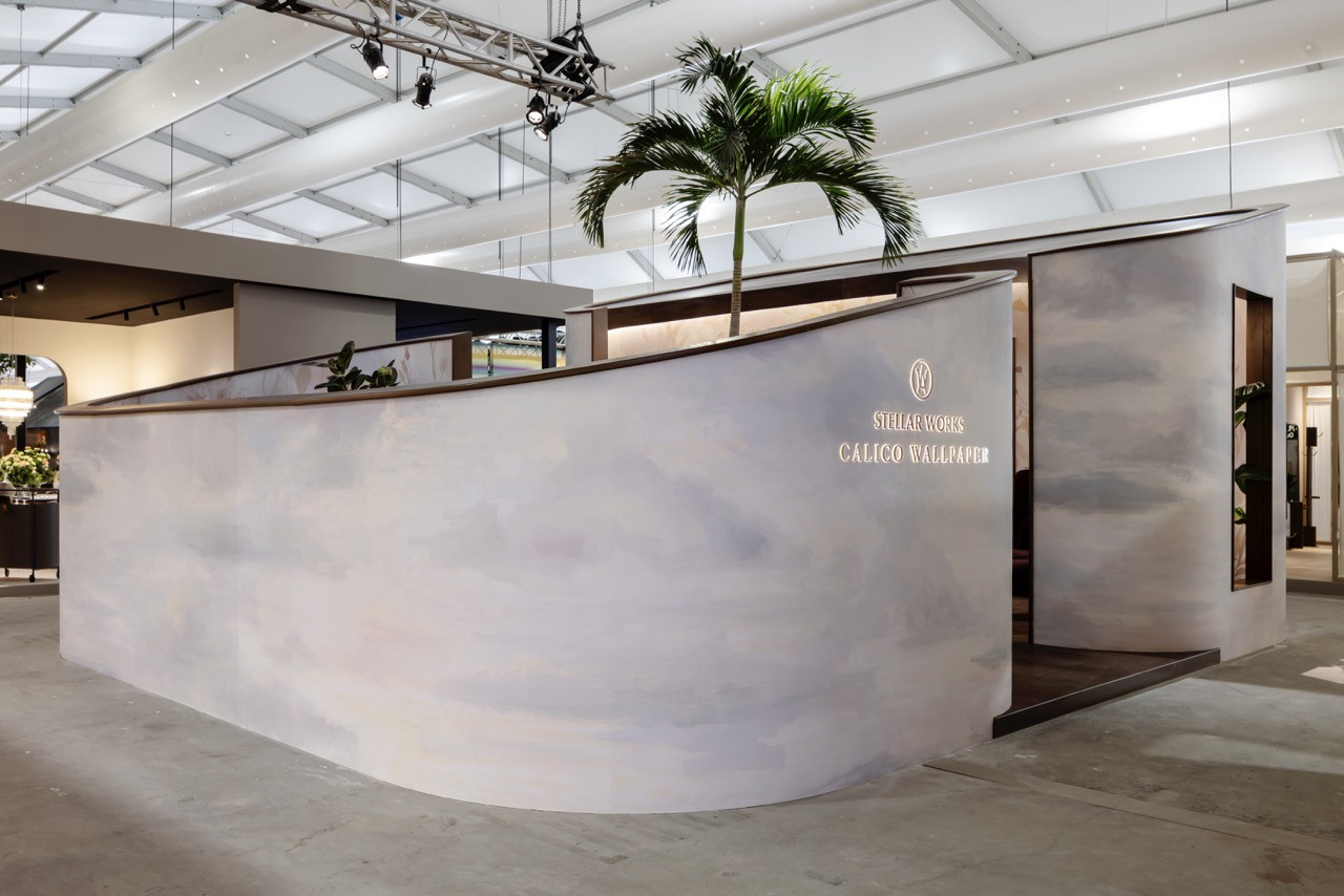 The booth entrance features a curved reception desk clad in Calico Wallpaper's Atmosphere collection with its impressionistic cloud pattern and soft gradient finish