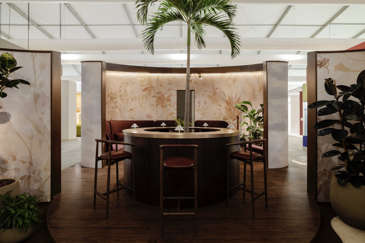 The bar area anchors Omar Al Gurg's booth design with a circular counter, Charlie Collection bar stools by Yabu Pushelberg, and a live palm tree overhead, backed by Calico Wallpaper’s Arcadia.