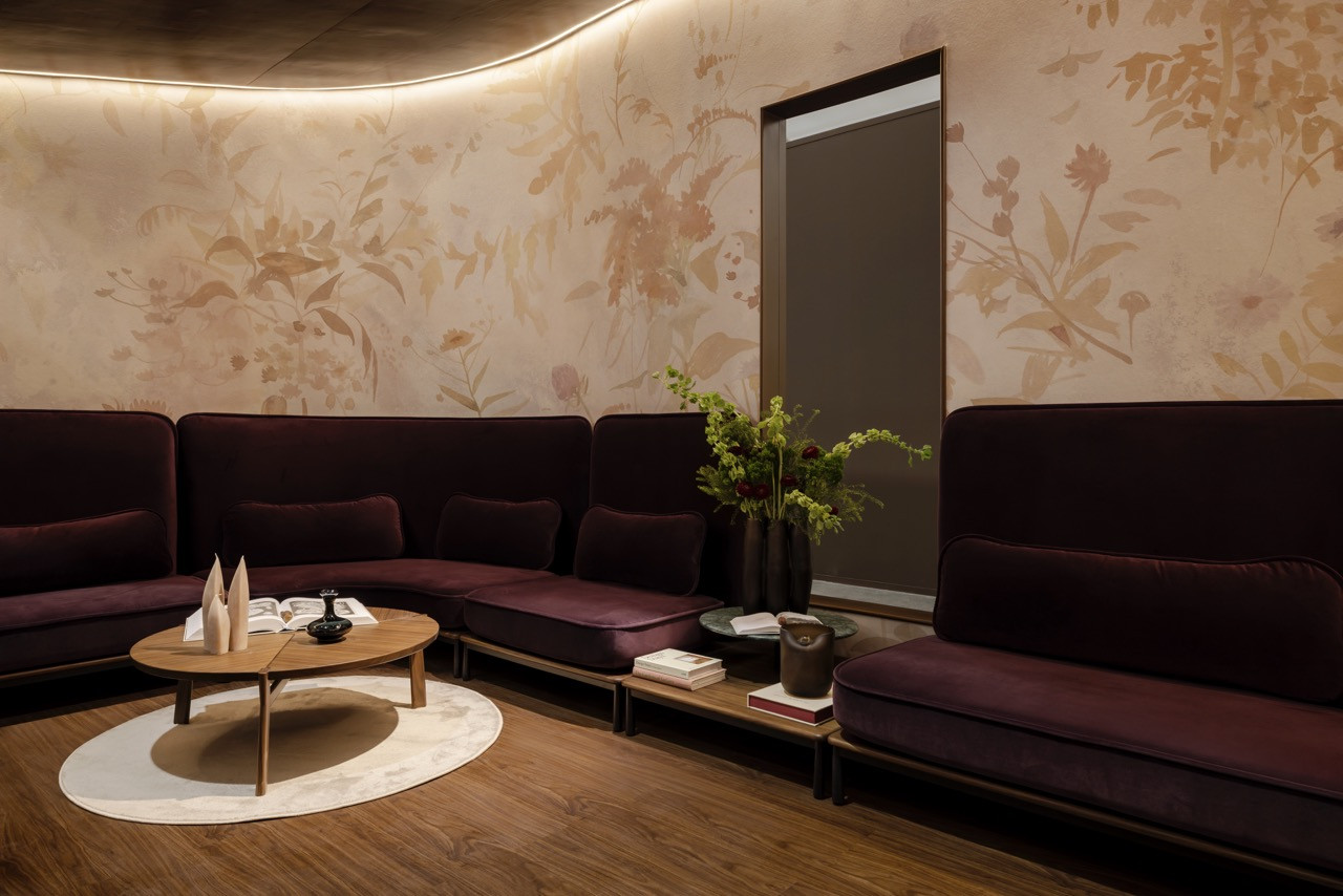 The lounge area features aubergine velvet seating by Stellar Works against Calico Wallpaper’s Arcadia collection, depicting botanical motifs in muted beige and gold tones.