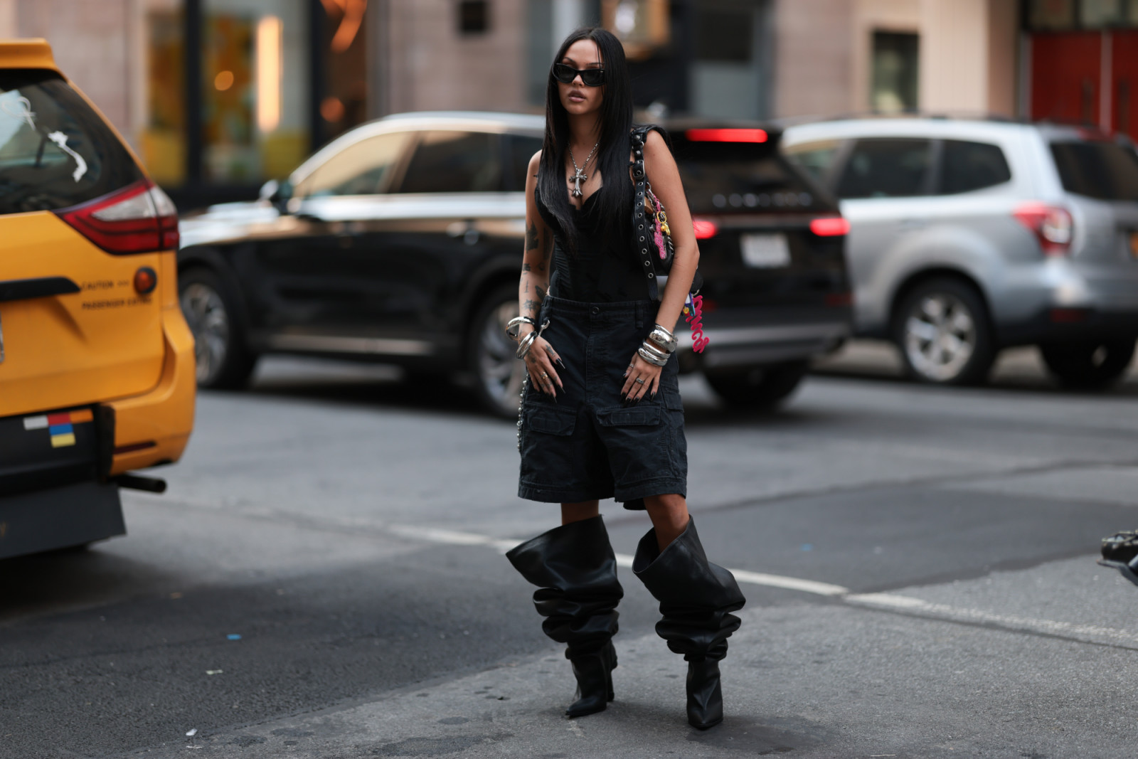 NEW YORK, NEW YORK - SEPTEMBER 14: A guest seen wearing a narrow cat-eye black sunglasses and a heavy silver cross pendant necklace, giving the look a gothic undertone, a black corset-style top, structured and body-hugging, layered with a sleeveless studded vest that adds toughness, an oversized black cargo shorts, loose and masculine, creating contrast against the fitted top, Balenciaga le Cagole black leather bag with colorful keychains attached to the strap, stacked with chunky silver bangles, paired wit