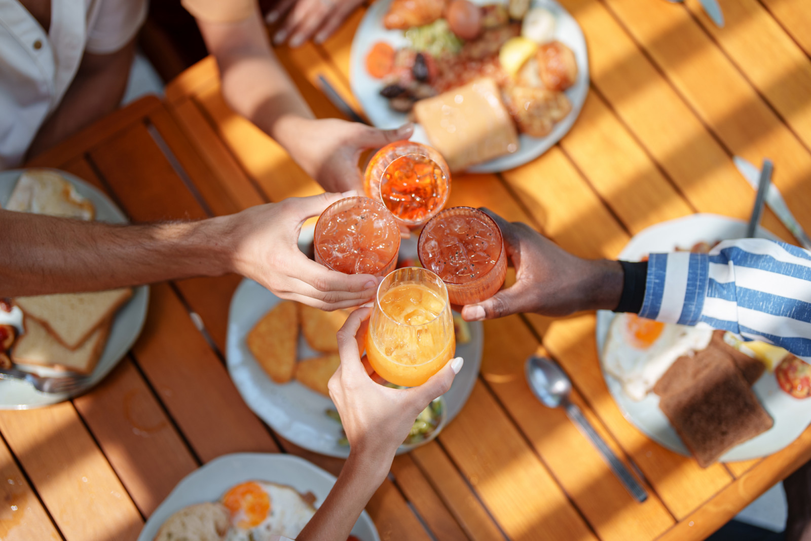 Close-up of diverse friends' hands raising glasses of fresh juice over a vibrant and nutritious breakfast spread on a wooden table, capturing the joy and togetherness of the moment.