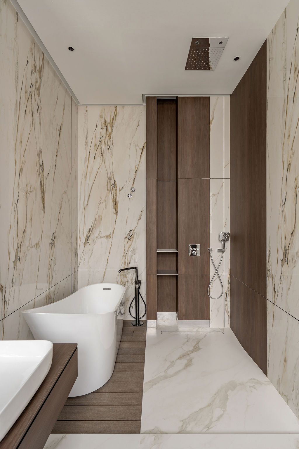 A freestanding tub sits on timber decking alongside marble walls and timber cabinetry with recessed storage