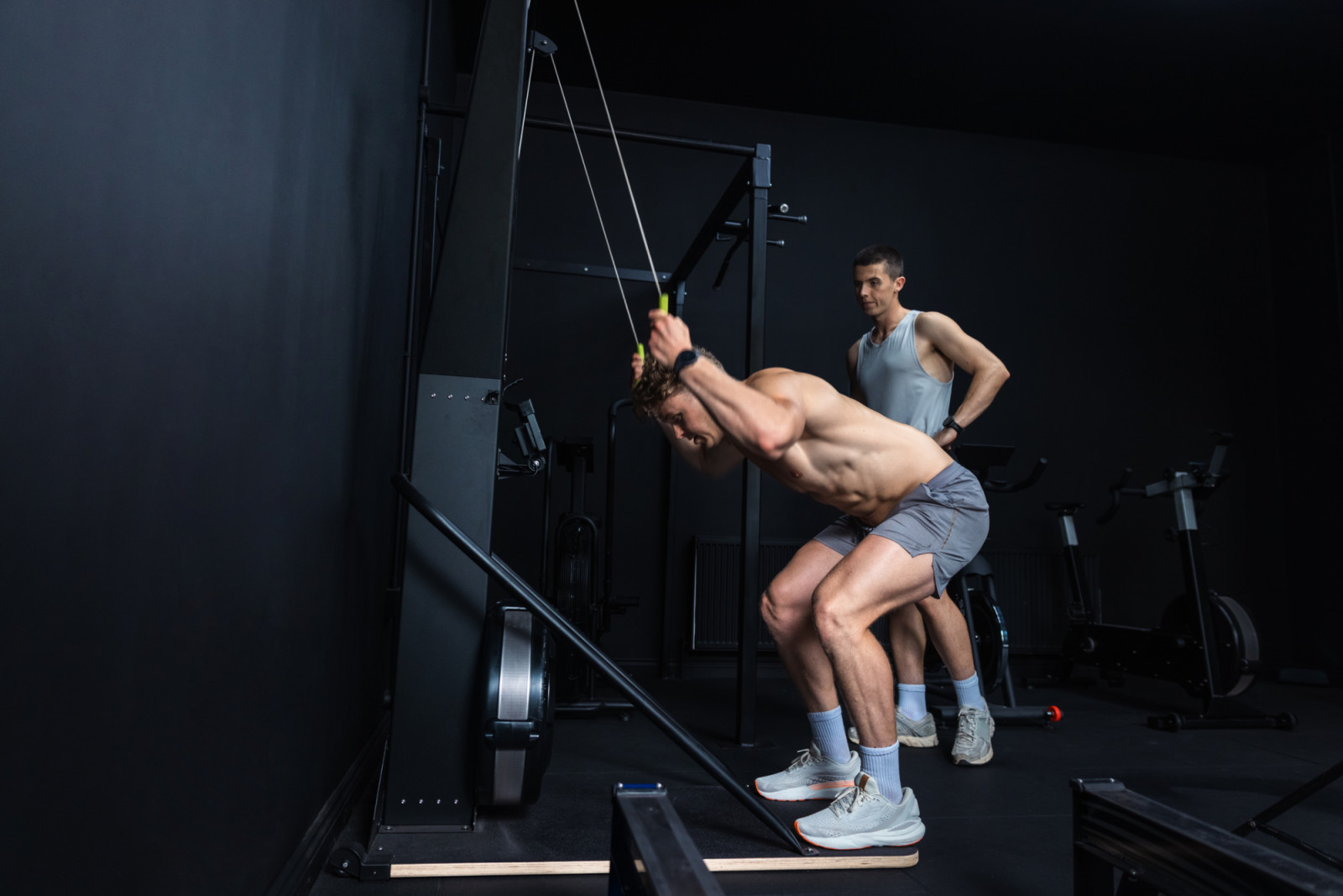A wide shot of two young men exercising with a SkiErg machine in a modern gym. One man is wearing grey shorts and sports shoes; he performs a resistance training movement targeting strength and muscle definition. The other man stands to the side and watches.
Videos are available similar to this scenario.